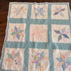 May include: A square quilt featuring a grid of nine white blocks, each containing a multi-colored star pattern made of various fabric scraps. The blocks are separated and bordered by a solid light blue fabric, creating a structured design.