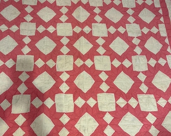 May include: A handmade quilt featuring a repeating pattern of white squares and diamonds on a pink backdrop. The quilt has a geometric design, with the white shapes creating a contrasting visual effect. The edges are finished with a white border.