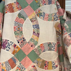 May include: A double wedding ring quilt featuring a white background with interlocking rings composed of various multi-colored floral, geometric, and polka dot fabric patches. The design includes solid pink and sage green square accents.