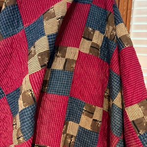May include: A patchwork quilt featuring a repeating pattern of square fabric blocks. The design consists of solid magenta squares, blue squares with a white grid pattern, and tan squares with thin dark stripes and small floral motifs.