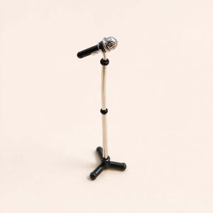 May include: Miniature microphone and stand. The microphone has a silver head and a black handle. The stand is silver with black adjustment rings and a black tripod base. The background is a solid, light beige color.