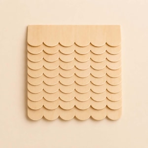 May include: A square, light-colored wooden decorative piece with a repeating scallop pattern. The piece is made of multiple wooden elements arranged in rows and columns, creating a textured surface. The background is a solid, light beige color.
