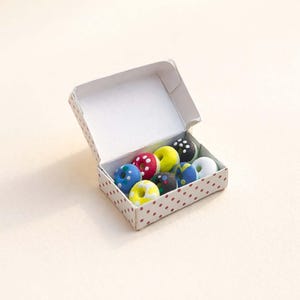 May include: A box of miniature, colorful donuts with various toppings, including polka dots and sprinkles. The donuts are arranged inside a white box with a red polka dot pattern.