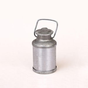 Peut inclure: Une petite cruche à lait en métal de style vintage, avec un couvercle à charnière et une poignée en fil de fer. La cruche a un corps cylindrique et une base circulaire. Le métal a un aspect légèrement vieilli. Le design est simple et fonctionnel.