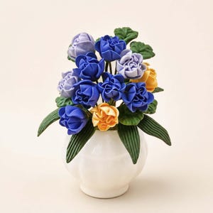 Puede incluir: Un jarr&oacute;n de cer&aacute;mica blanca contiene un ramo de flores artificiales. El arreglo presenta rosas azules, amarillas y lavanda con hojas verdes. El jarr&oacute;n tiene forma redondeada con una amplia abertura.