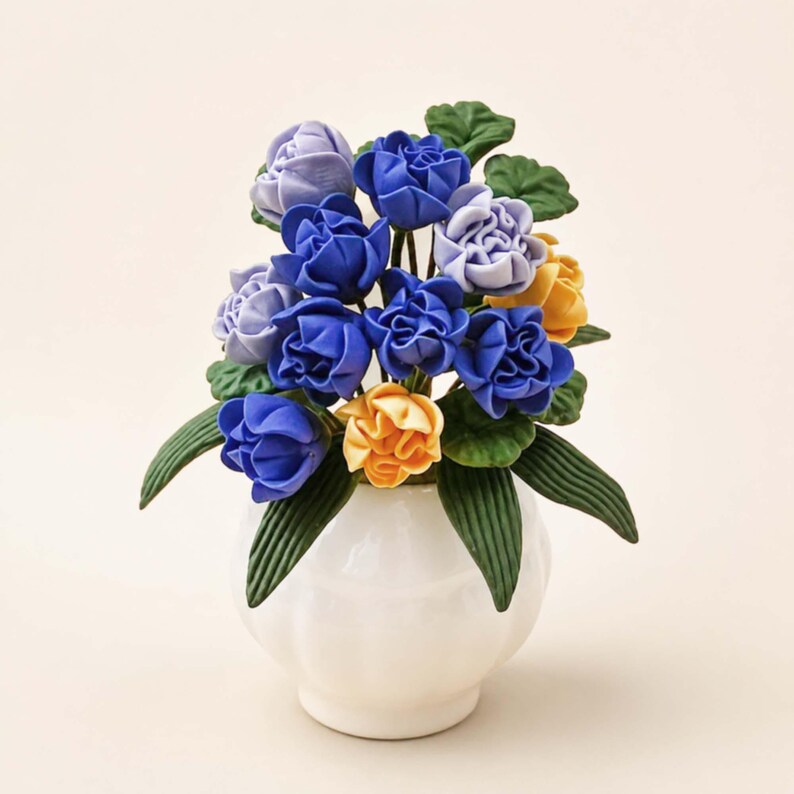 Puede incluir: Un jarr&oacute;n de cer&aacute;mica blanca contiene un ramo de flores artificiales. El arreglo presenta rosas azules, amarillas y lavanda con hojas verdes. El jarr&oacute;n tiene forma redondeada con una amplia abertura.