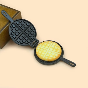 May include: A black waffle iron with a waffle inside. The waffle iron is open and the waffle is visible. The waffle iron has a handle on one side.