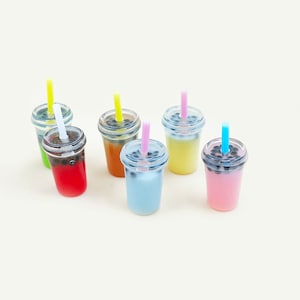 Puede incluir: Seis vasos de plástico en miniatura con pajitas llenas de líquidos de diferentes colores y bolas de tapioca negras. Los vasos son verde, naranja, amarillo, azul, rosa y rojo.
