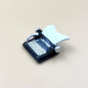 Puede incluir: Una máquina de escribir en miniatura de color negro con un teclado plateado y un portapapeles blanco. La máquina de escribir está hecha de plástico y tiene aproximadamente el tamaño de un juguete pequeño.