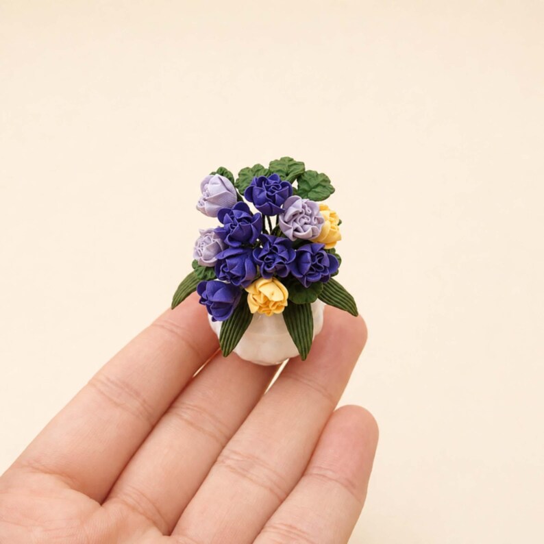 Puede incluir: Un arreglo floral en miniatura en una maceta blanca. El arreglo presenta flores moradas, amarillas y lavanda con hojas verdes. Las flores est&aacute;n dispuestas en una peque&ntilde;a maceta de cer&aacute;mica blanca, creando una est&eacute;tica encantadora y artesanal.