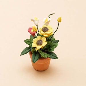 Könnte beinhalten: Ein kleiner Terrakotta-Topf gefüllt mit handgefertigten Tonblumen. Das Arrangement umfasst gelbe Sonnenblumen, weiße Calla-Lilien und eine kleine rosa Blume, alle umgeben von grünen Blättern. Der Topf steht vor einem weichen, neutralen Hintergrund.