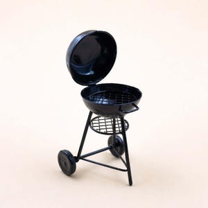 以下が含まれることがあります： 蓋が開いたミニチュアの黒い炭火焼きグリル。グリルは、金属製の格子、下の棚、2つの車輪を備えた丸い調理面を備えています。グリルは金属製で、光沢のある仕上がりです。