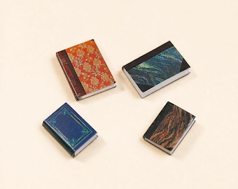 May include: Four miniature books with detailed covers. One book has a red floral pattern, another has a blue cover with gold accents, and the other two have abstract designs in shades of brown and blue. The books are arranged on a cream-colored surface.