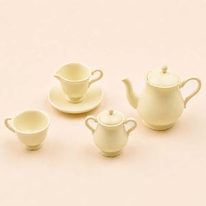 Peut inclure: Un service à thé en céramique crème, comprenant une théière, un sucrier, une crémière, une tasse et une soucoupe. L'ensemble a un design classique avec des formes arrondies et des poignées délicates.