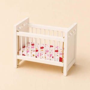 Puede incluir: Una cuna de madera blanca en miniatura con barras verticales y detalles metálicos dorados. La cuna presenta un colchón con un estampado de fresas rosas y lunares dorados sobre fondo blanco.