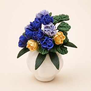 Puede incluir: Un jarr&oacute;n de cer&aacute;mica blanco contiene un ramo de flores artificiales. El arreglo presenta rosas azules, amarillas y lavanda con hojas verdes. El jarr&oacute;n tiene una forma redondeada con un borde festoneado, creando una exhibici&oacute;n floral cl&aacute;sica.