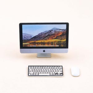 Op de afbeelding: Een miniatuur computeropstelling met een zilver-zwarte monitor met een berglandschap. Een zwart-wit toetsenbord en een witte muis bevinden zich onder de monitor. Het Apple-logo is zichtbaar op de monitor.