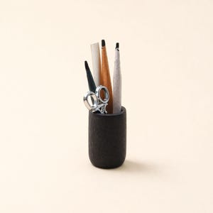 May include: A small, dark wooden pencil holder containing several pencils, a small pair of scissors, and a white paper tube. The pencils are in various colors, including black, brown, and gray. The scissors are silver.