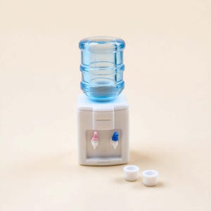 Puede incluir: Un pequeño dispensador de agua blanco con una botella de agua azul transparente en la parte superior. El dispensador tiene boquillas rosas y azules y dos tazas blancas pequeñas. El fondo es de color beige claro liso.