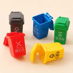 Op de afbeelding: Een set van vijf miniatuur plastic afvalbakken in zwart, blauw, groen, rood en geel. Elke bak is voorzien van een scharnierend deksel en specifieke iconen en tekst voor afvalcategorieën die op de voorzijde zijn gedrukt.