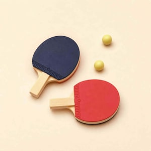 May include: Two table tennis bats, one navy blue and one red, with light wood handles, and two yellow table tennis balls. The bats have a black border against a cream background.