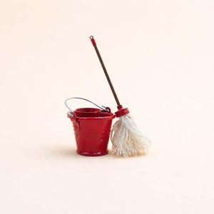 Può includere: Un piccolo secchio rosso con manico argentato e un mocio con manico marrone e testa bianca. Gli articoli per la pulizia sono su uno sfondo color crema. Il mocio è appoggiato al secchio.