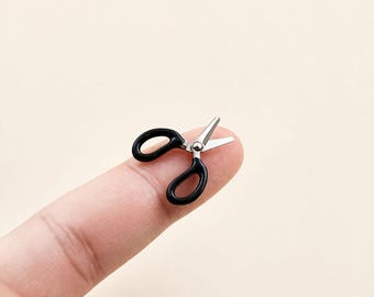 May include: Miniature black scissors with silver blades, resting on a fingertip. The small scissors have a classic design with looped handles. The blades are open, suggesting they are ready for use. The background is a soft, neutral color.