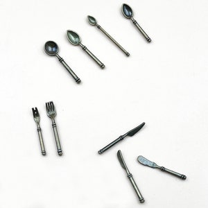 Puede incluir: Un juego de cubiertos en miniatura de color plateado, que incluye cucharas, tenedores y cuchillos. Los cubiertos están dispuestos sobre una superficie blanca.