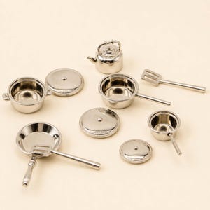 Puede incluir: Un juego de cocina en miniatura de color plateado que consta de una pequeña tetera, dos ollas con tapas a juego, una sartén y dos espátulas ranuradas. Cada pieza está fabricada con un material metálico pulido con un acabado superficial brillante.