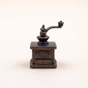 May include: A small, bronze-toned coffee grinder with a hand crank. The grinder features a square base with the word "CAFE" inscribed on the front. The crank is positioned at the top of the grinder.