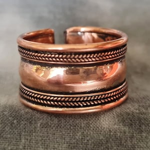 Pode incluir: Um anel de cobre com um design trançado em torno da parte superior e inferior. O anel é largo e tem uma forma arredondada.