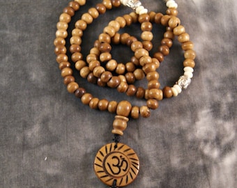 May include: A brown wooden mala bead necklace with a carved wooden Om pendant and silver accents. The necklace has a tassel with three strands of brown wooden beads and silver accents.