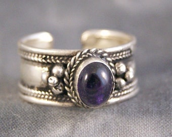 Può includere: Un anello d'argento con una pietra preziosa ametista incastonata in una fascia decorativa. La fascia presenta un design intrecciato e piccole perline.