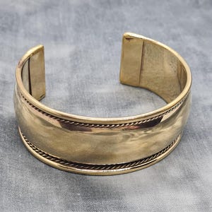 Può includere: Un braccialetto rigido in metallo dorato con una superficie liscia e arrotondata e un design a corda attorno al bordo.