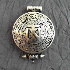 May include: Silver pendant with intricate filigree design and a central symbol. The symbol is surrounded by a circle of Tibetan script.