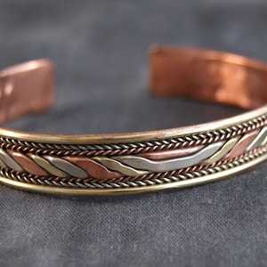 Könnte beinhalten: Ein Kupferarmband mit einem geflochtenen Design in Kupfer, Silber und Gold.