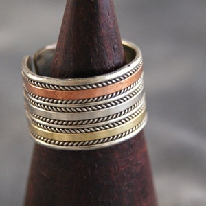 Puede incluir: Un anillo de plata con múltiples bandas, cada una con un diseño diferente. Las bandas están hechas de plata, cobre y latón. El anillo está en un expositor de madera.