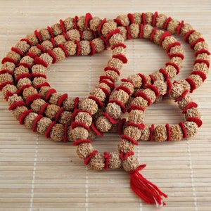 Puede incluir: Un collar de cuentas mala marrón y rojo con un borla. Las cuentas son de madera y tienen un hilo rojo envuelto alrededor.
