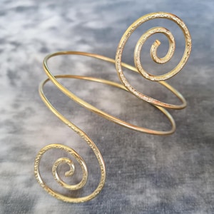 Può includere: Un bracciale dorato con un design a spirale. Il bracciale si avvolge attorno al braccio con due spirali decorative. Il metallo ha una finitura strutturata e il design è elegante. Un accessorio di moda.