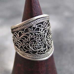 Può includere: Un anello color argento con un intricato design a filigrana. L'anello ha una fascia larga con un motivo dettagliato di curve e anelli, ed è esposto su un cono di legno marrone scuro.