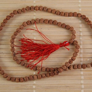 May include: A brown wooden mala bead necklace with a red tassel. The necklace is laid out on a bamboo mat.