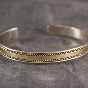 Puede incluir: Un brazalete de plata con una banda dorada y un diseño de cuerda.