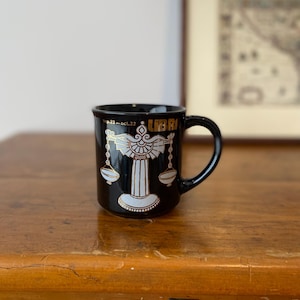 Pode incluir: Caneca de cer&acirc;mica preta com o s&iacute;mbolo do signo de Libra em dourado e branco. A caneca tem uma al&ccedil;a preta e a palavra "LIBRA" &eacute; impressa em dourado. As datas "sep.23-oct.23" tamb&eacute;m s&atilde;o impressas.