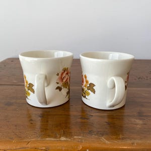 May include: Two white ceramic mugs with floral designs. The mugs have a brown, yellow, and pink floral pattern. The mugs are on a wooden surface.