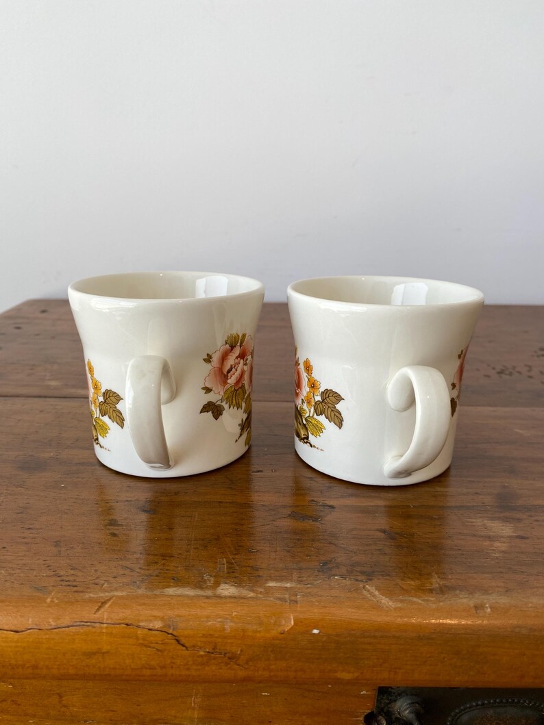 May include: Two white ceramic mugs with floral designs. The mugs have a brown, yellow, and pink floral pattern. The mugs are on a wooden surface.