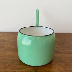 May include: A mint green enamel saucepan with a long handle and a black rim. The interior is white. The saucepan is sitting on a wooden surface. The handle has a hole for hanging.