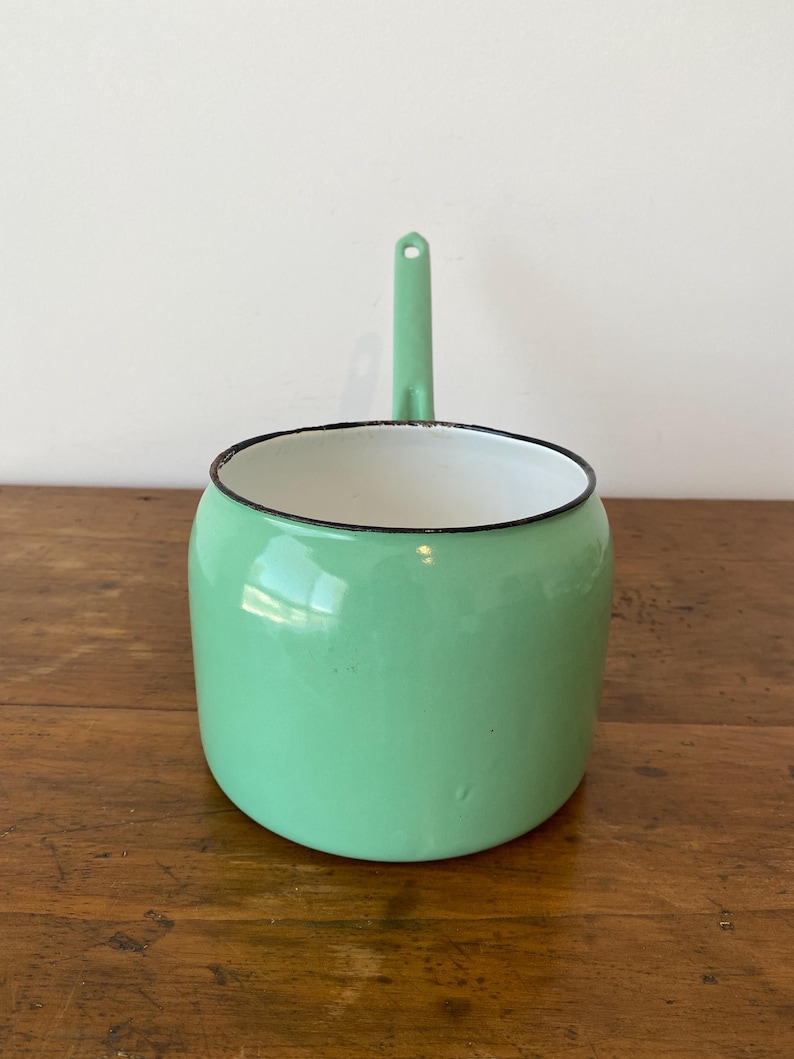 May include: A mint green enamel saucepan with a long handle and a black rim. The interior is white. The saucepan is sitting on a wooden surface. The handle has a hole for hanging.