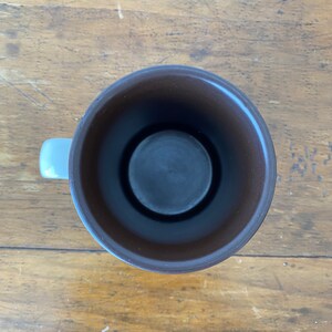 May include: A brown ceramic mug with a white handle. The mug is empty and the inside is visible.
