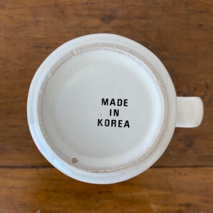 May include: White ceramic mug with a black inscription that reads "MADE IN KOREA" on the bottom.