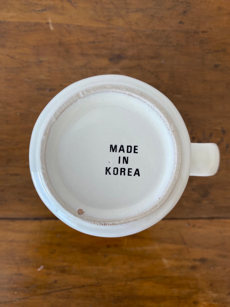 May include: White ceramic mug with a black inscription that reads "MADE IN KOREA" on the bottom.